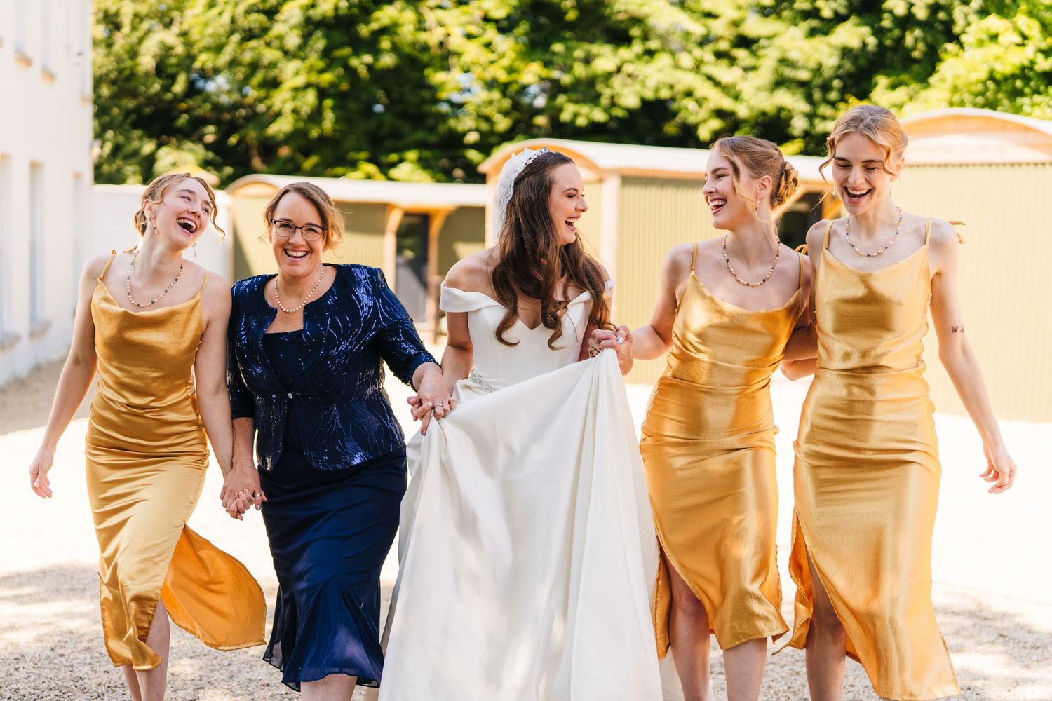 Poulaphouca Wedding photograA wedding party walks together outdoors with bridesmaids wearing elegant gold silk dresses and one in navy blue.
