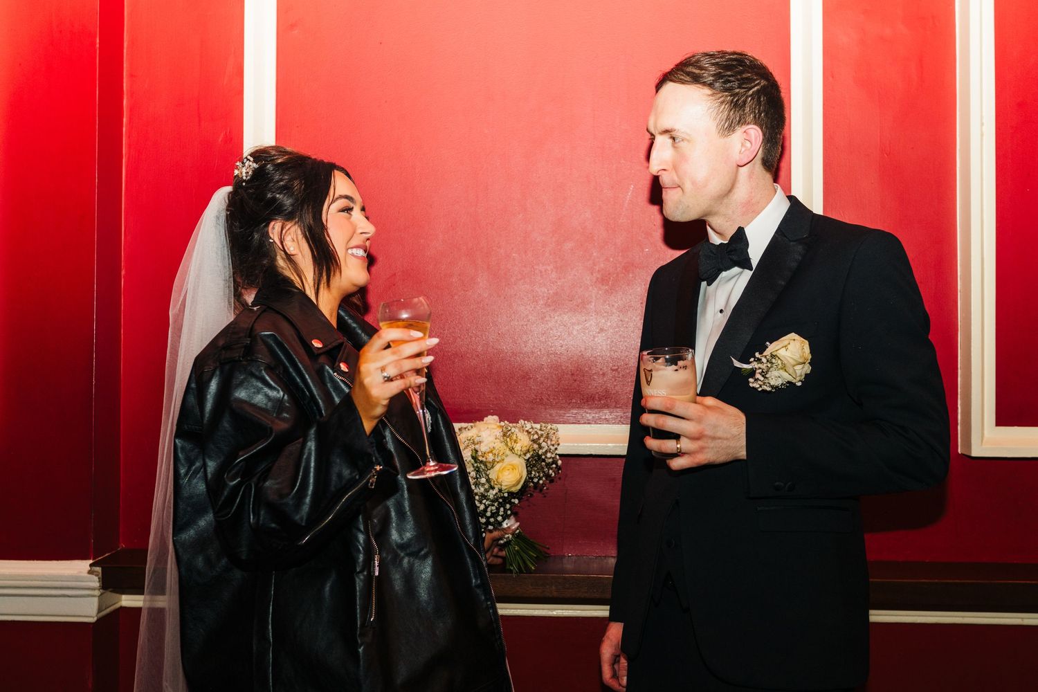 A couple enjoys drinks together against a vibrant red wall during their wedding celebration. Wedding PHOTOGRAPHER Dublin