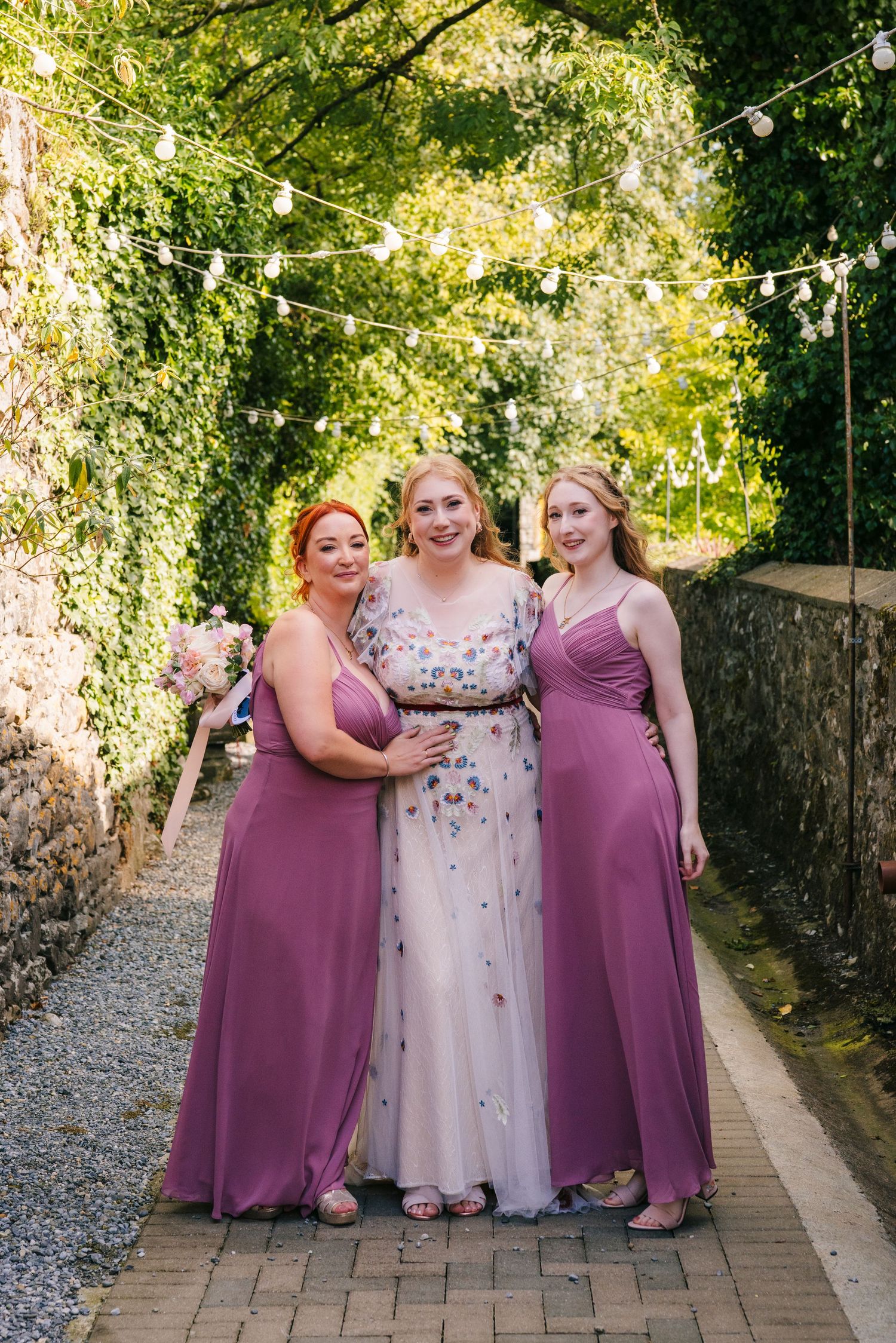 Three people in formal attire pose on a garden path with string lights overhead and lush greenery in the background. . Village Barn Tyrrells