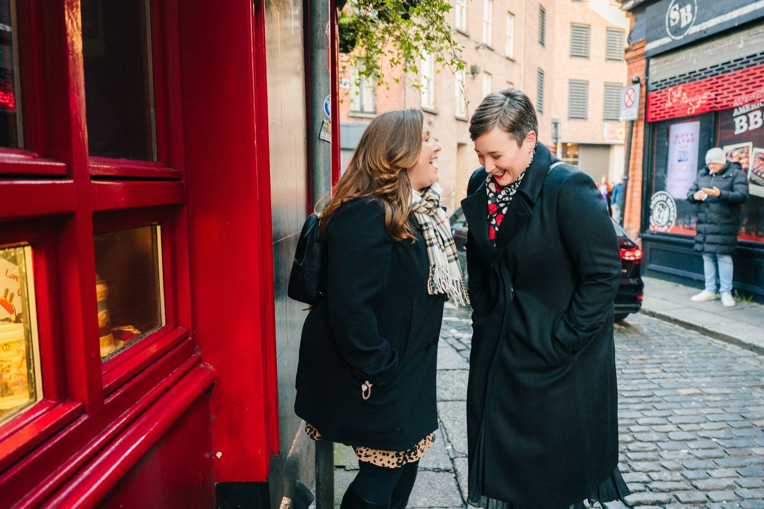 A couple shares a romantic moment on a cobblestone street lined with red storefronts in Dublin's Temple Bar district. Family walking through