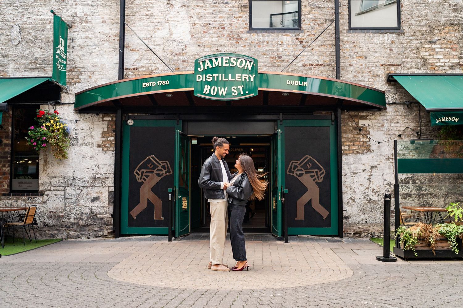 A brick building exterior with green storefront signage and stone arches forms a panoramic view of historic architecture. Couple's Photoshoo
