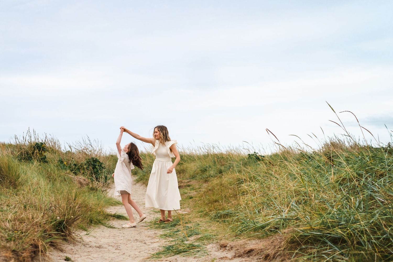 A bride in a flowing white dress climbs a grassy sand dune path near the beach.  Family photographer Dublin Ireland