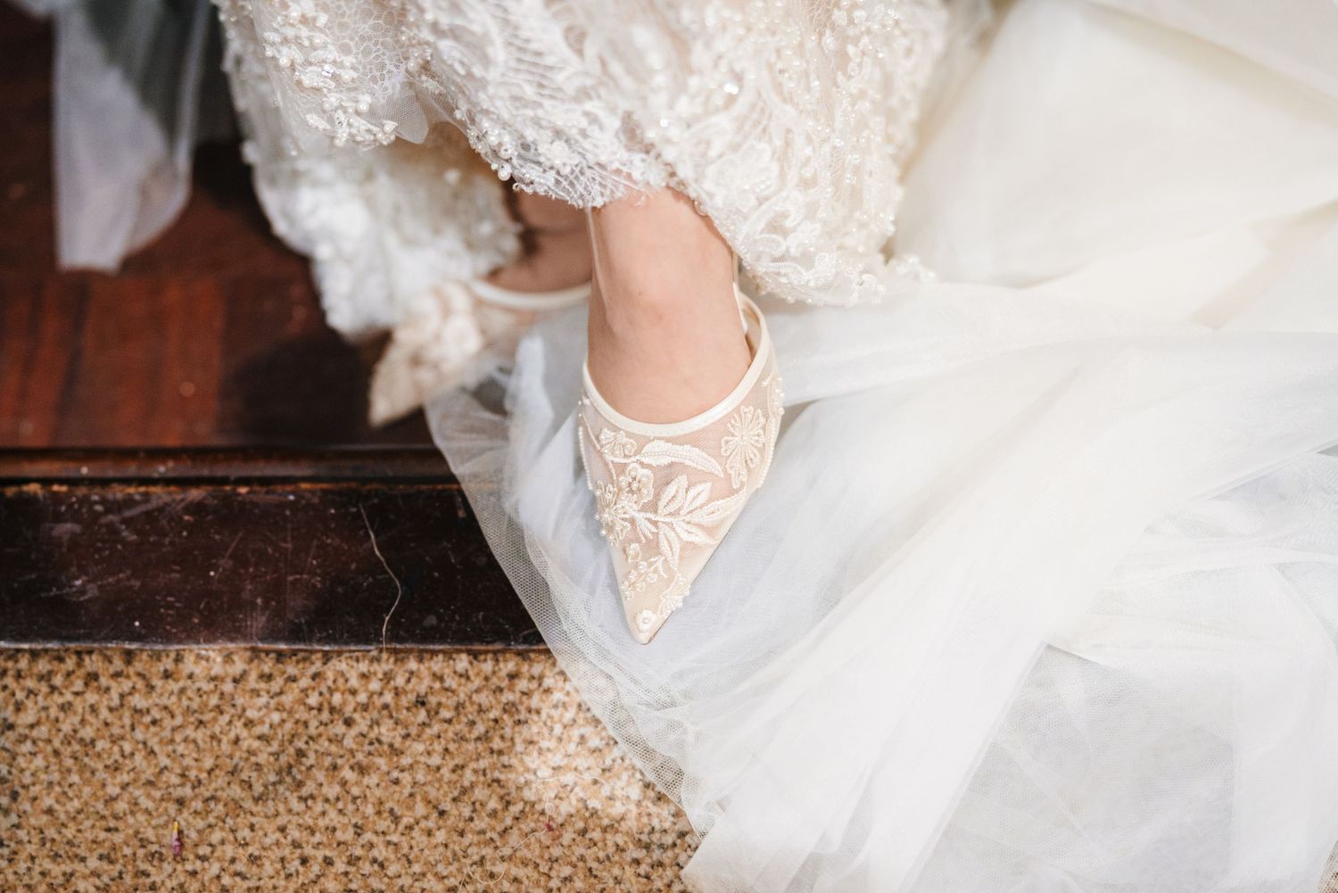 Close-up details of a white lace wedding dress and shoes against a textured floor.