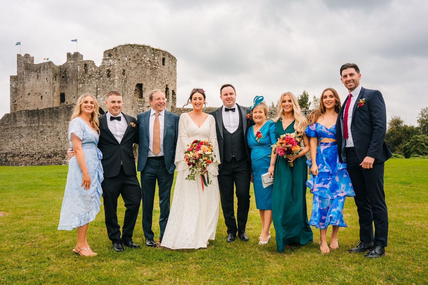 A wedding party poses together on grass in front of a historic castle ruins on an overcast day. Wedding Photography Trim Castle Ireland