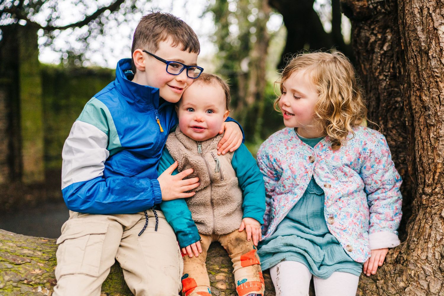 Three children sitting together on a log in a forest setting, wearing casual autumn clothing and smiling. Family photographer Dublin Ireland