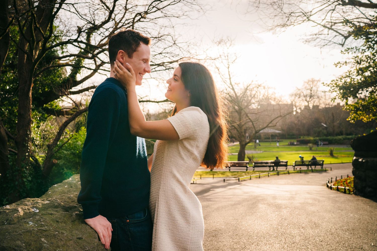 A loving couple embraces in a sunlit park with park benches and bare winter trees in the background. Surprise proposal dublin photography