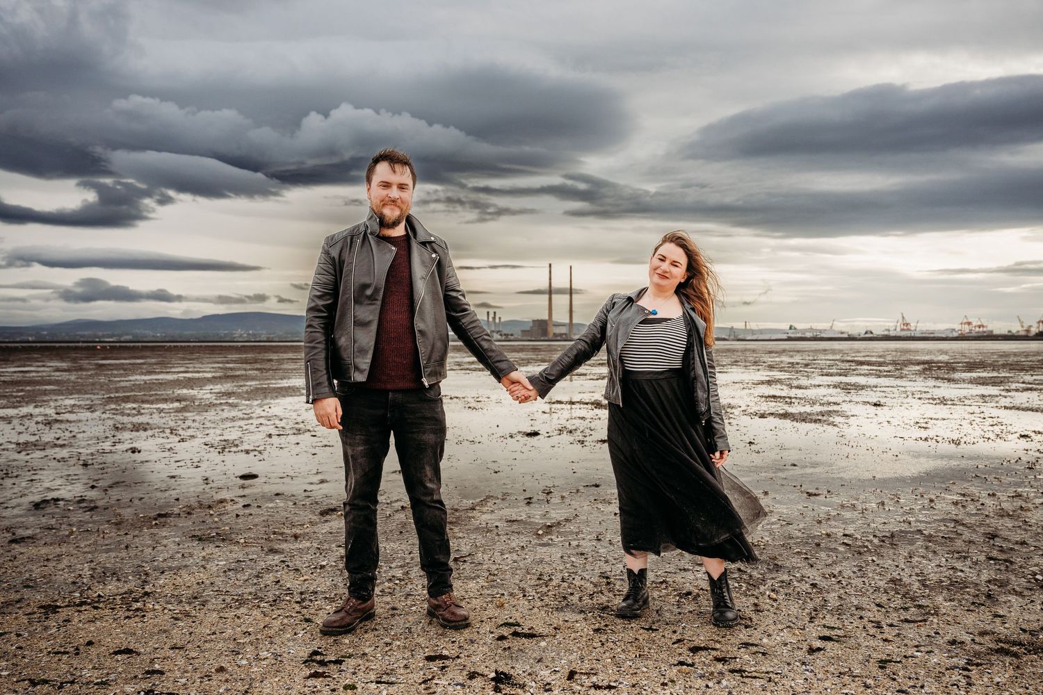 Playful couple poses on empty beach under dramatic storm clouds. engagement PHOTOGRAPHER Dublin