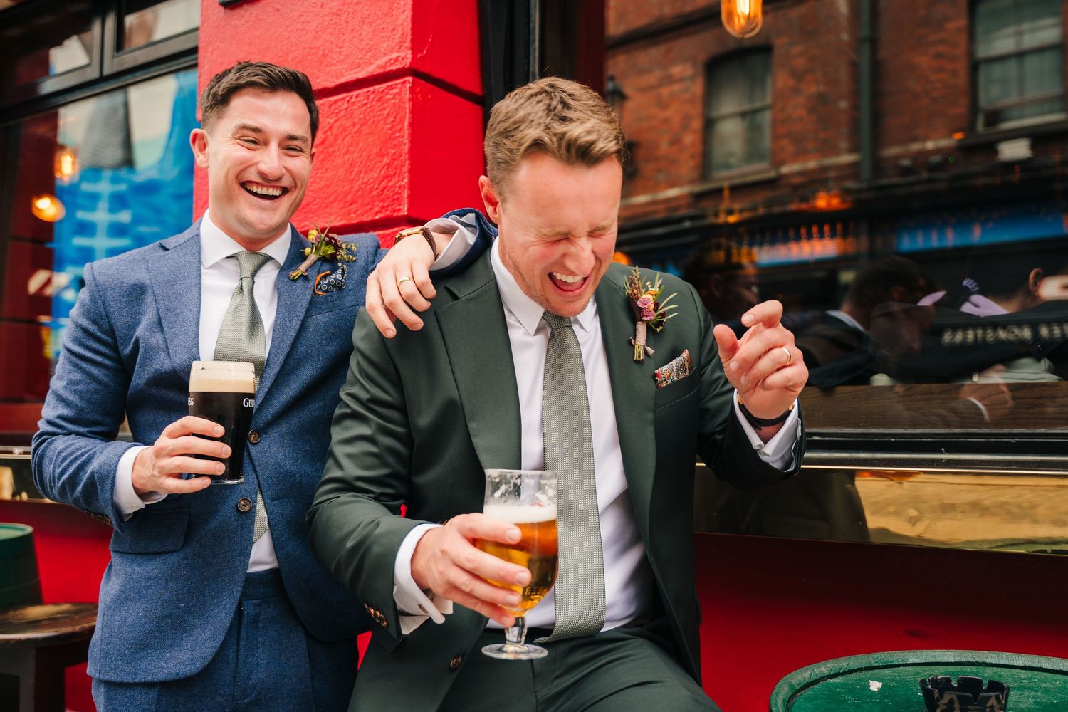 Two men in suits share a laugh while enjoying beers outside a restaurant with a red exterior wall. Same sex wedding PHOTOGRAPHER Dublin
