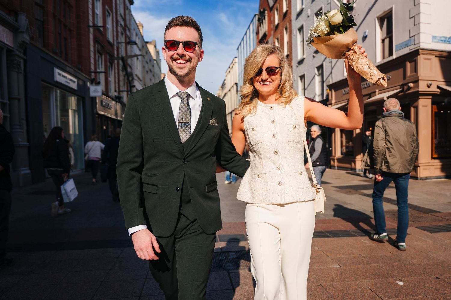 A couple walks joyfully down a city street, with the bride in white and groom in dark suit amid urban architecture. City Hall Dublin wedding