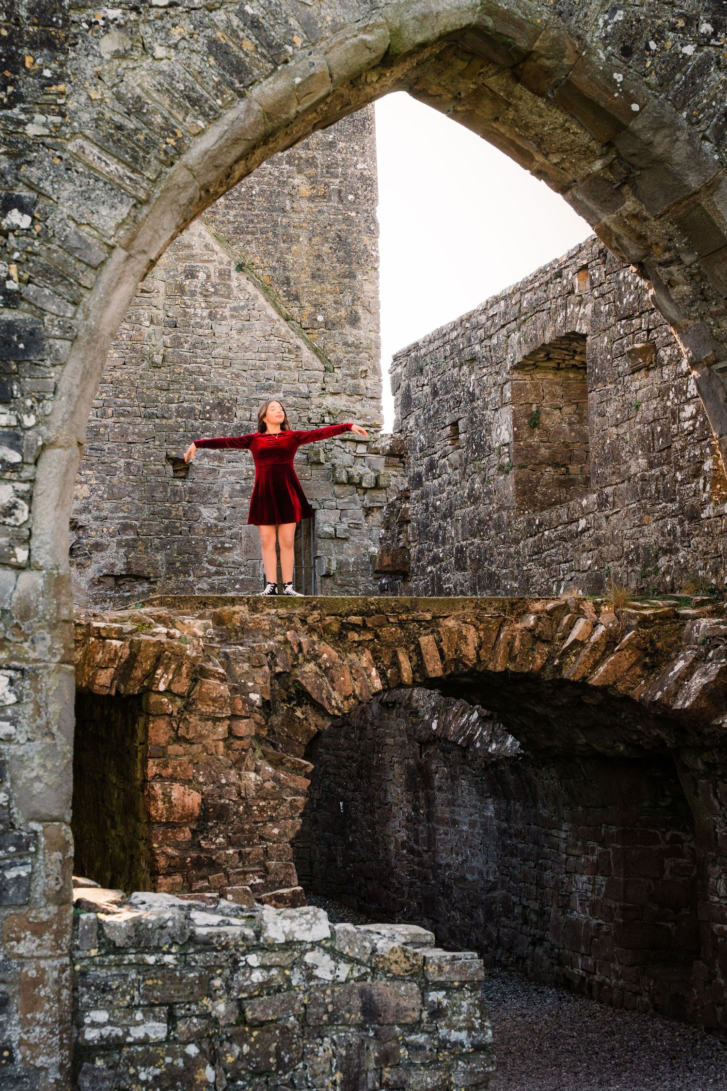 Figure in red dress stands with arms outstretched in ancient stone archway ruins. Family PHOTOGRAPHER Dublin