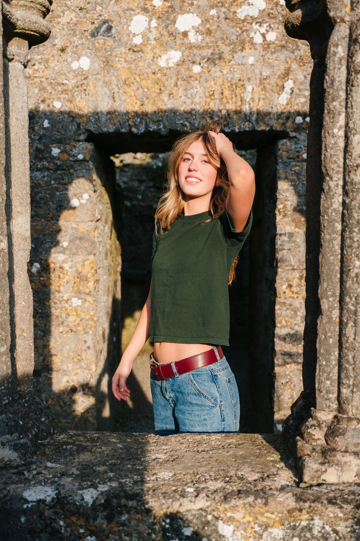 Sun-drenched figure in green crop top and jeans stands in rustic stone archway on bright summer day. Photographer Ireland dublin