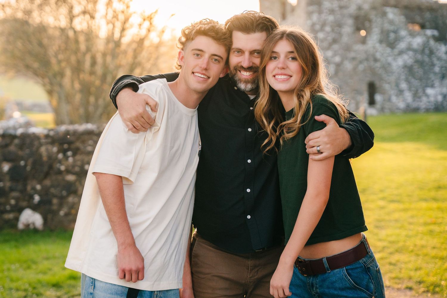 Three people embracing and laughing together outdoors during golden hour with stone walls visible in background. Photographer Ireland dublin