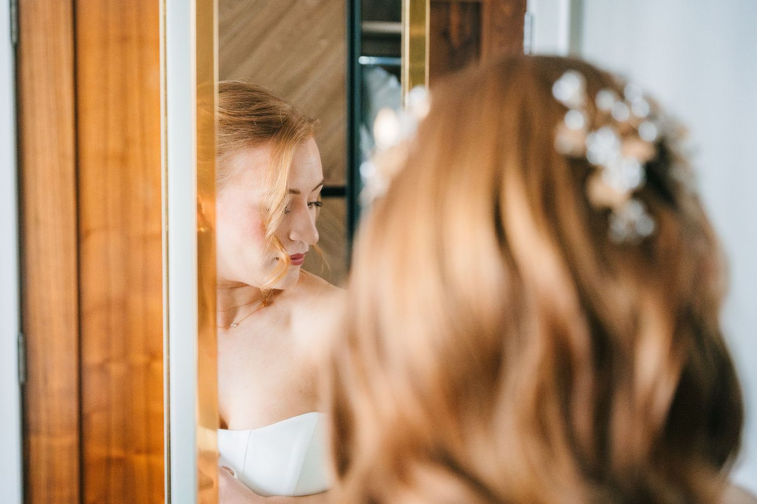 Bride in white dress looks in mirror while getting ready, with delicate floral hair accessories visible. Dublin wedding photogrAPher Bewleys