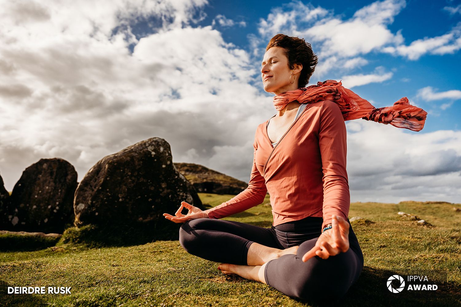 A person in coral attire meditating in lotus position against a dramatic cloudy landscape. PHOTOGRAPHER Dublin