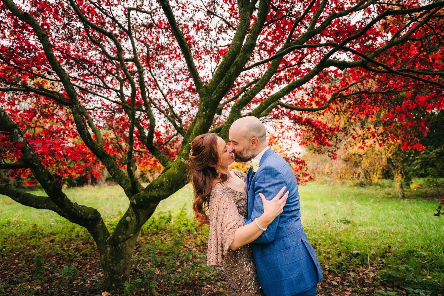 A romantic moment captured under a vibrant red Japanese maple tree in a lush garden setting during autumn season. Wedding PHOTOGRAPHER Dubli