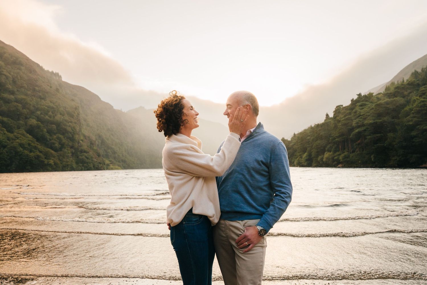 Two people share a tender moment at sunset by a lake surrounded by misty mountains. PHOTOGRAPHER DUBLIN IRELAND