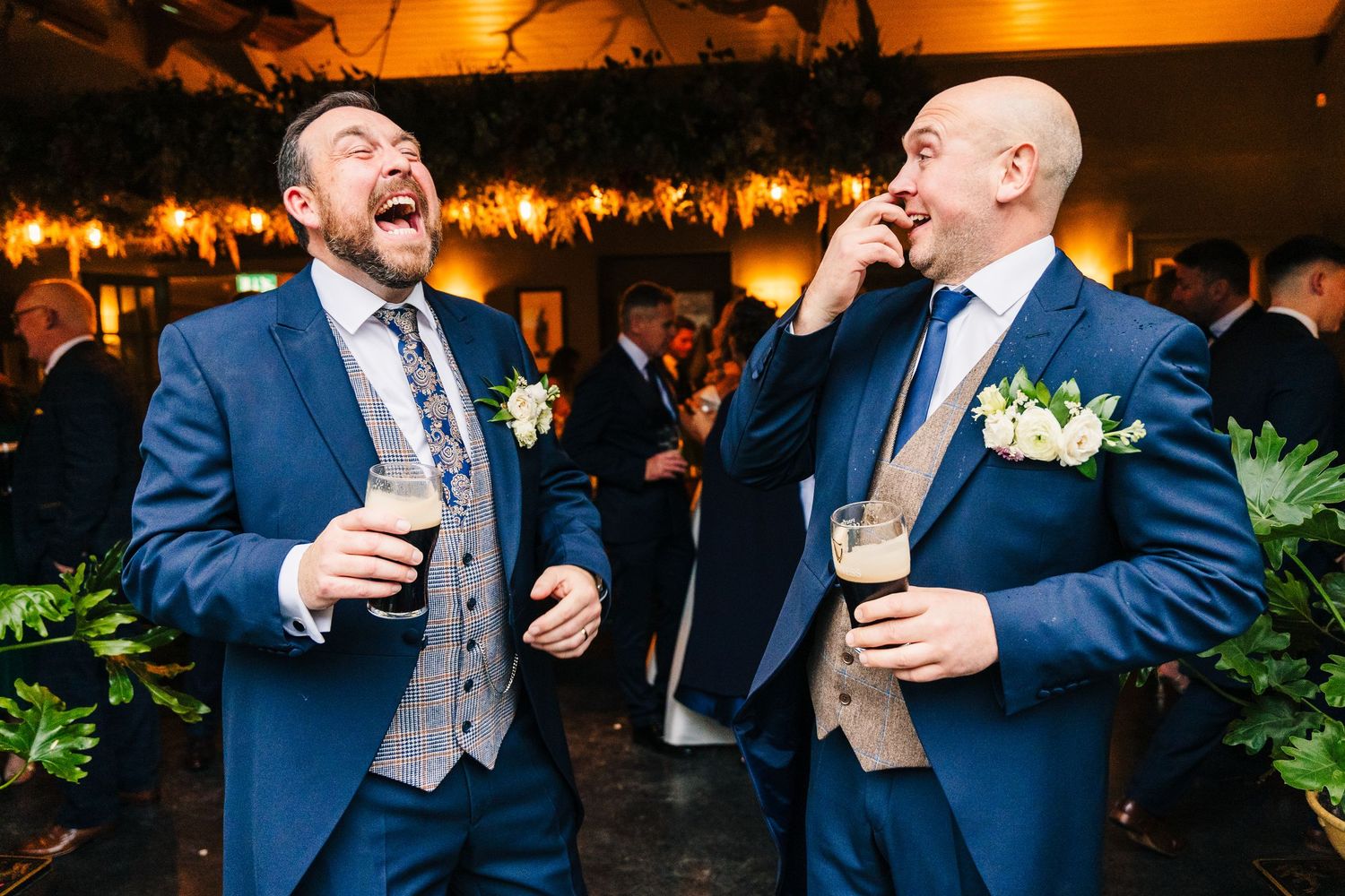 Groomsmen share laughter and drinks at an evening wedding reception with warm lighting.