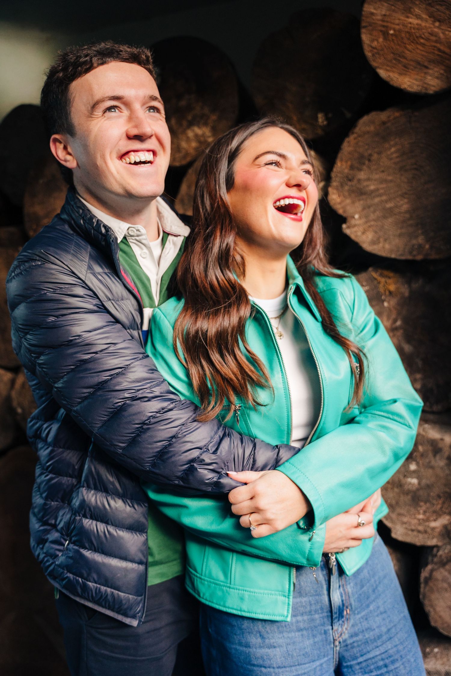 A couple laughing together while embracing in front of stacked logs, wearing a navy puffer jacket and turquoise leather jacket. engagement p