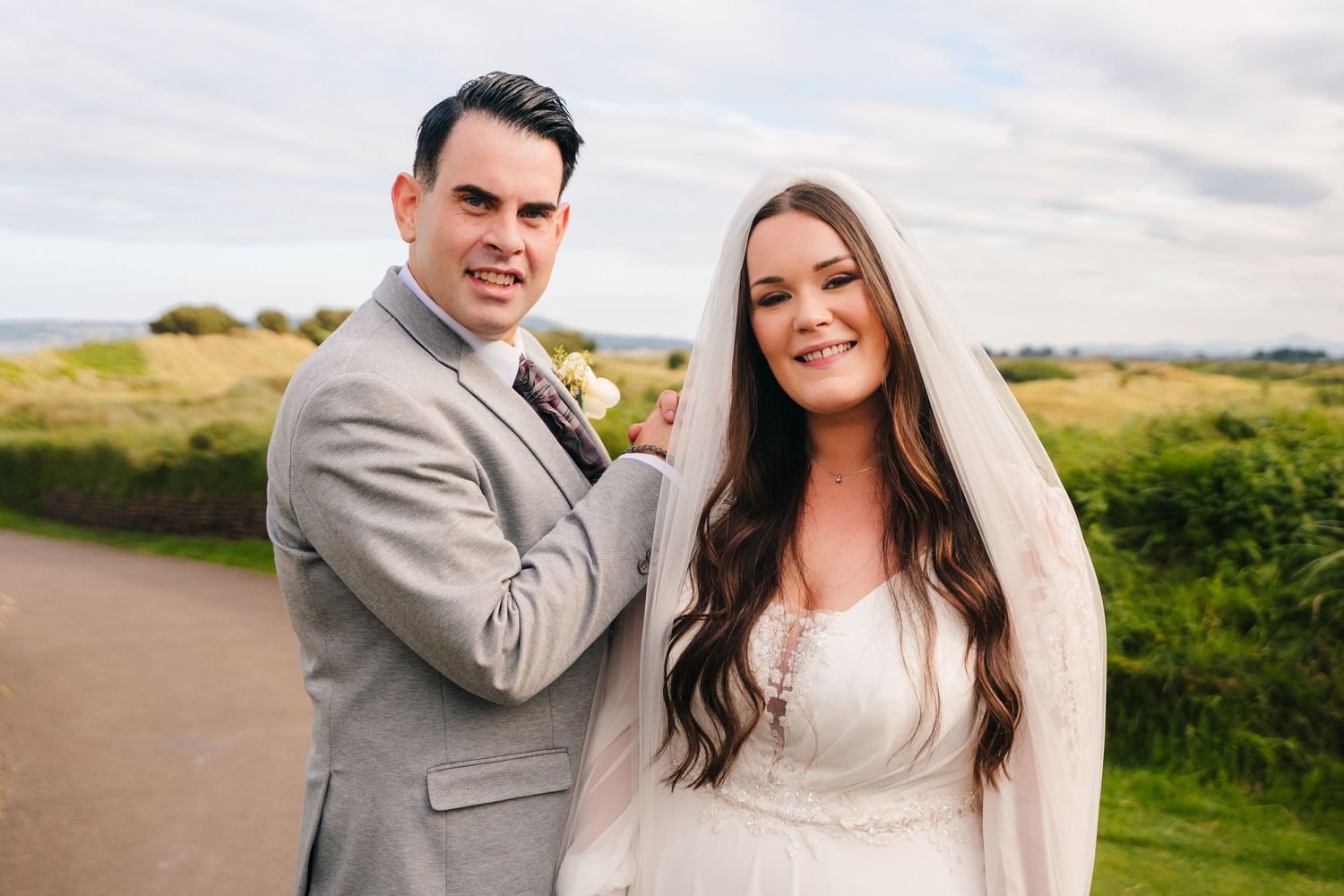 A couple in wedding attire poses together on a scenic path during their special day. Portmarnock Hotel Wedding PHOTOGRAPHER Dublin