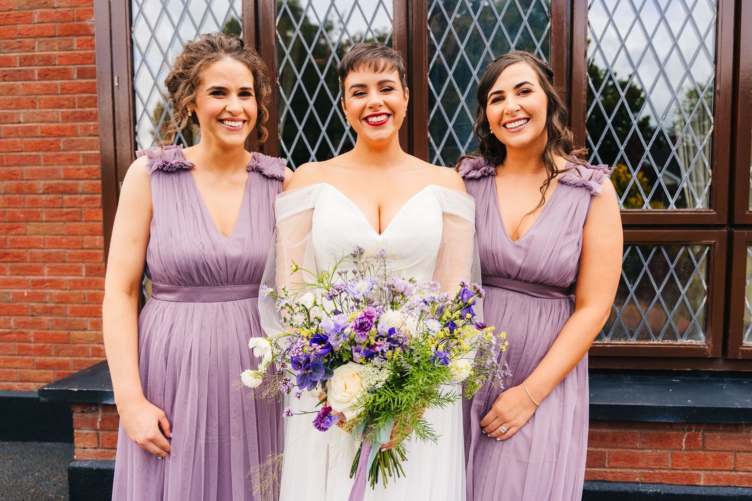 Wedding party members in lilac bridesmaid dresses pose with bouquets of purple and white wildflowers. Millhouse slane wedding photographer d