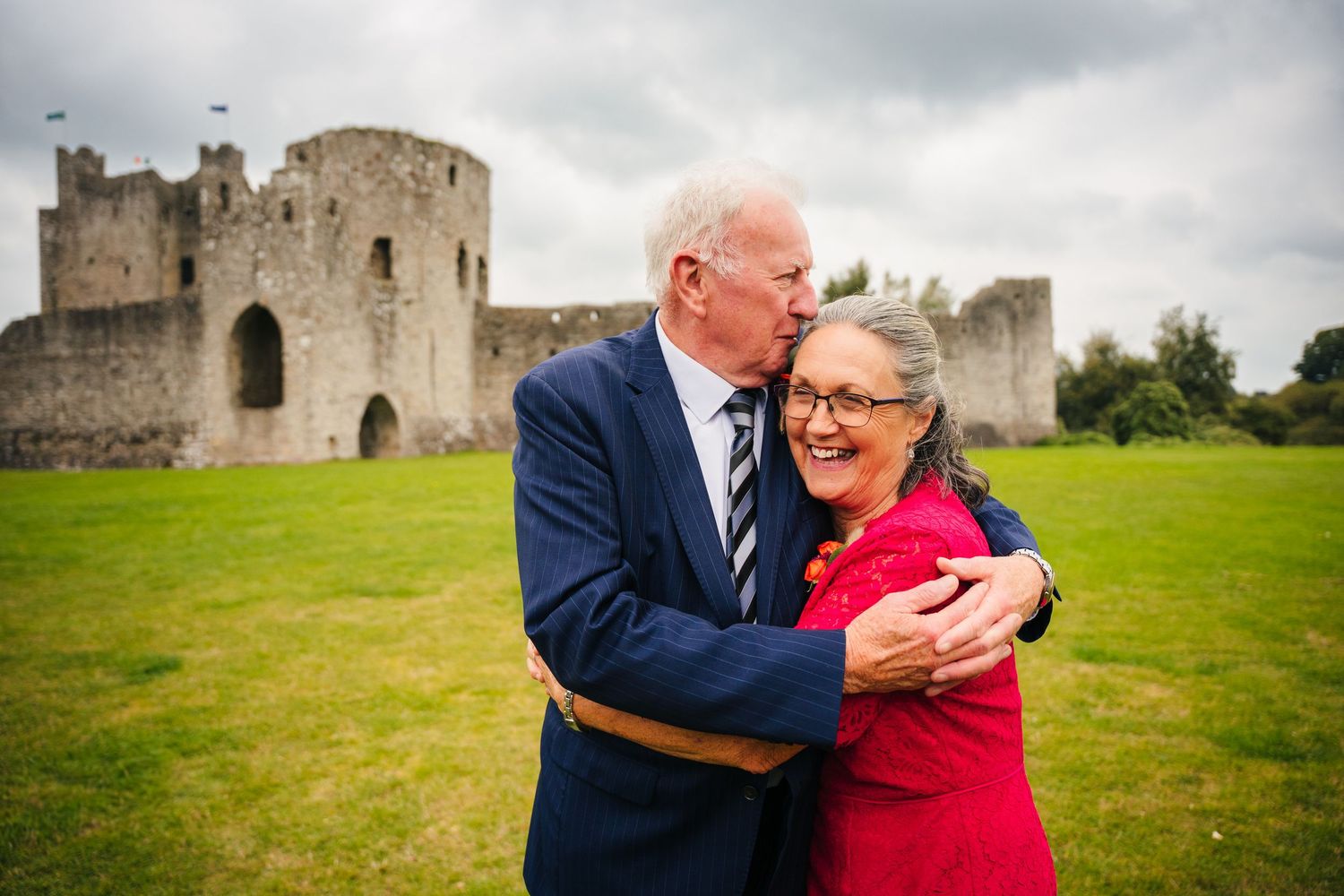 A couple embraces on the grounds of a historic castle under cloudy skies. Wedding Photography Trim Castle Ireland