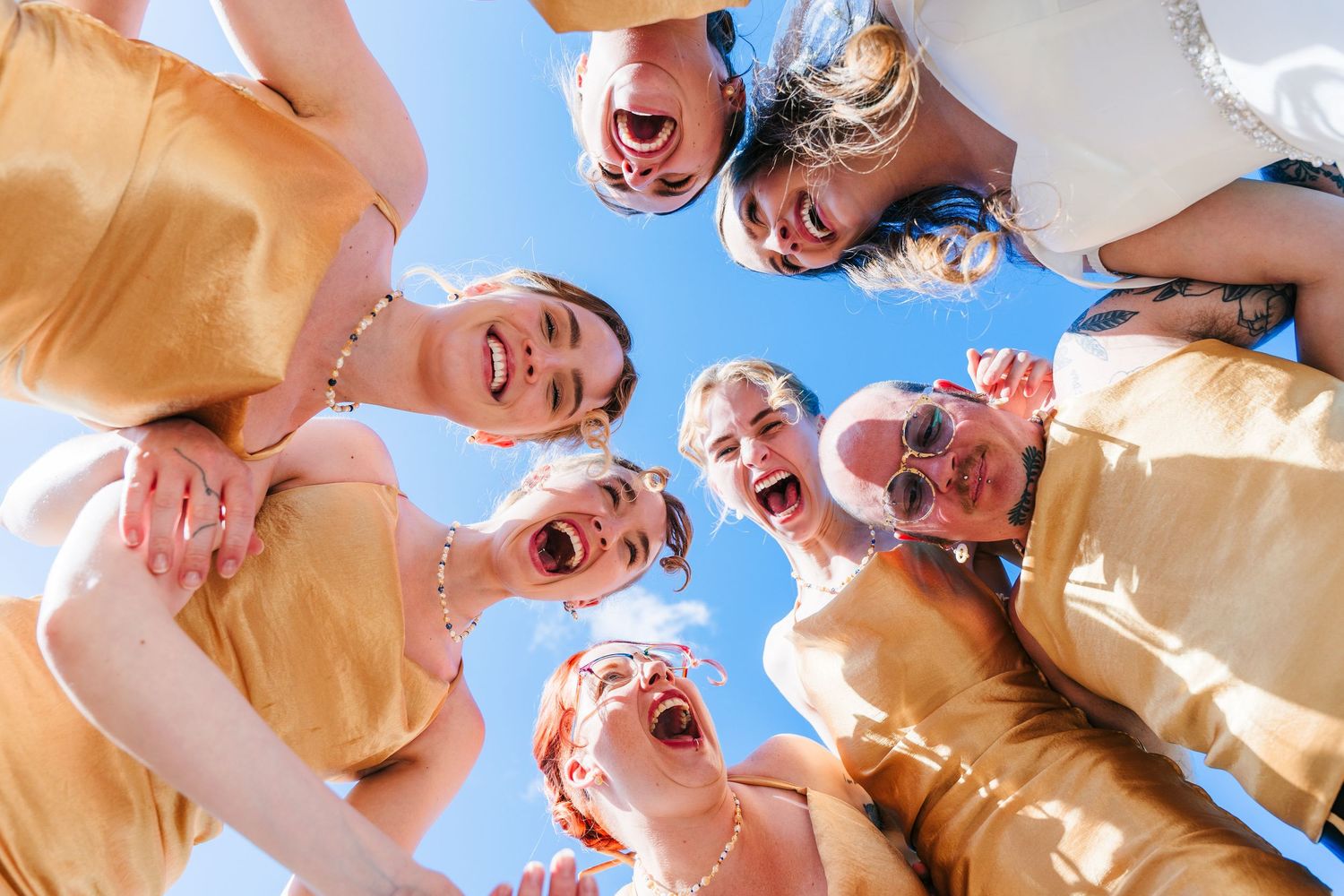 Poulaphouca Wedding photographer wicklow  Circle of people in golden bridesmaid dresses looking down at camera against bright blue sky.