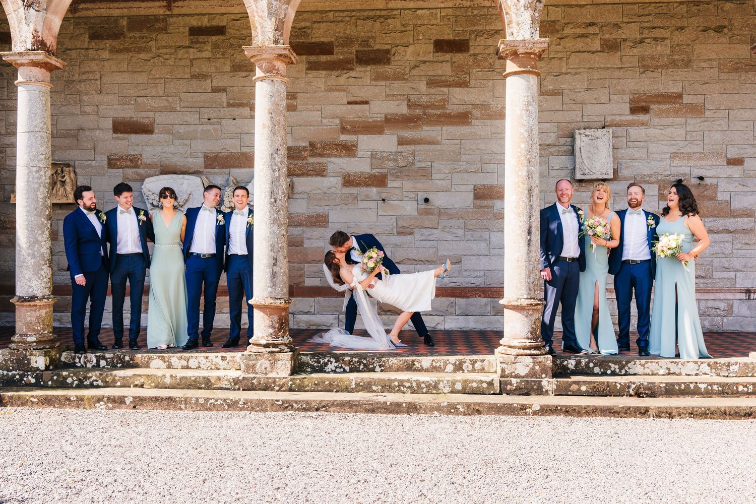 Wedding party poses on stone veranda with ornate columns and brick walls during outdoor celebration. Castle Leslie Wedding Photographer Dubl