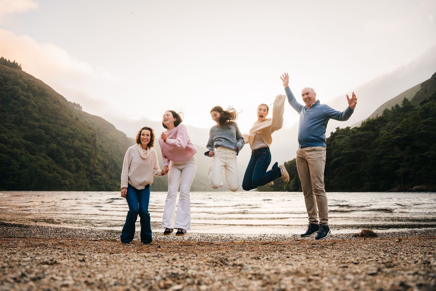 Family photographer Ireland Dublin Joyful family jumping together on a beach with mountains in the background during sunset.