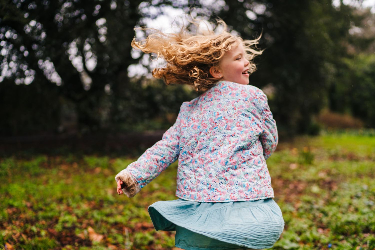 Blonde hair flowing in the wind as a child in a floral jacket spins outdoors on green grass. Family photographer Dublin Ireland