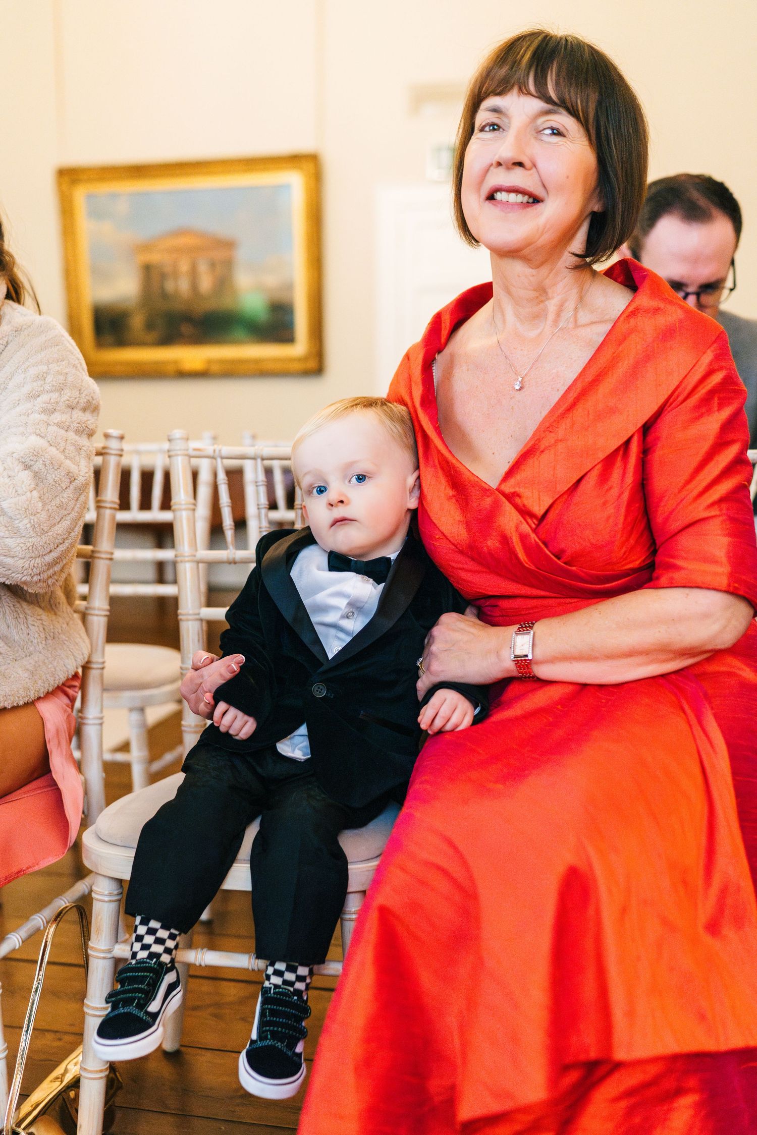 A baby in a black outfit sits on a person's lap wearing a vibrant coral red dress during a formal event.  Wedding PHOTOGRAPHER Dublin