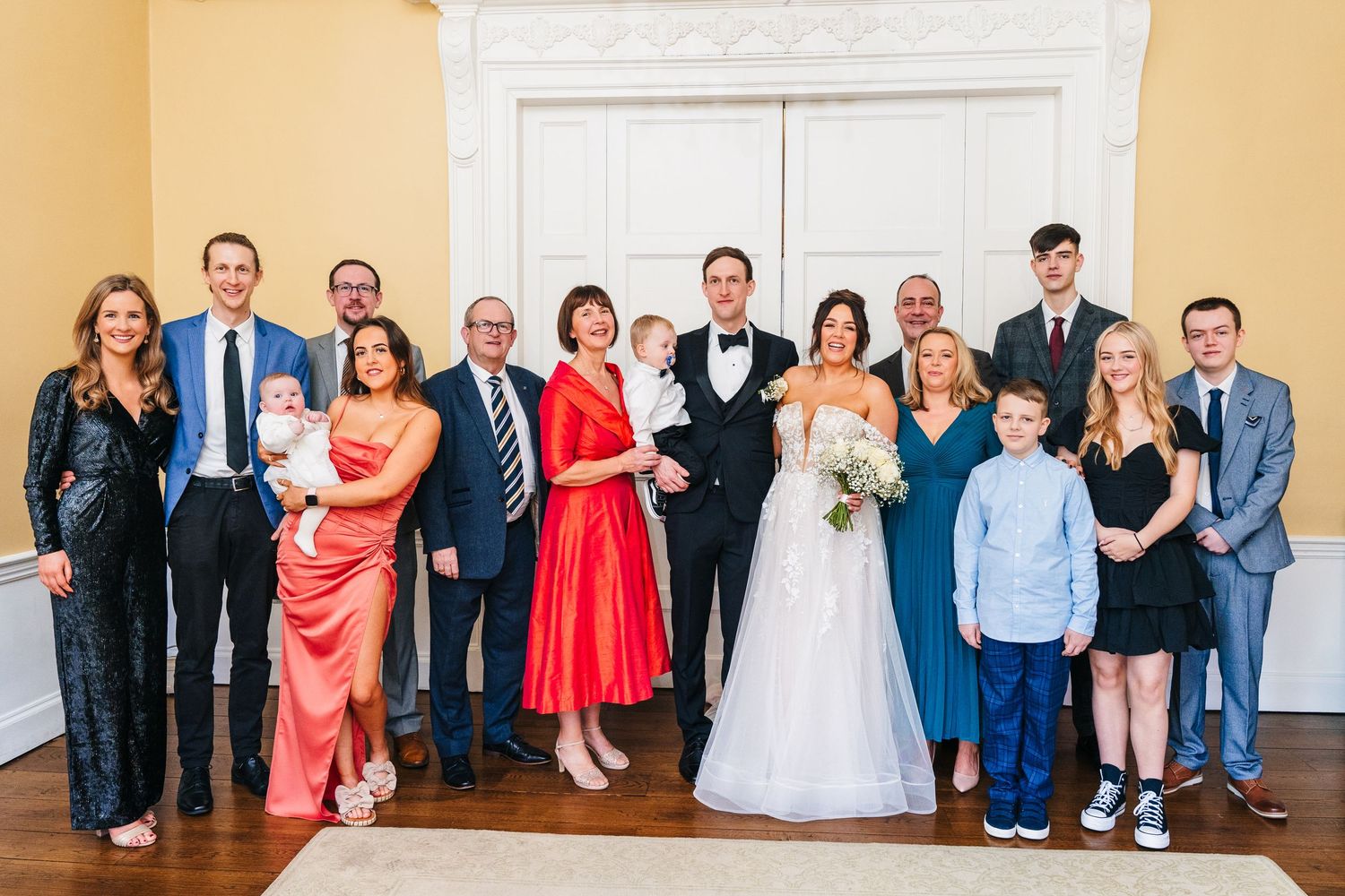 A large family wedding party poses together for a formal group photo in front of an ornate white wall. Wedding PHOTOGRAPHER Dublin