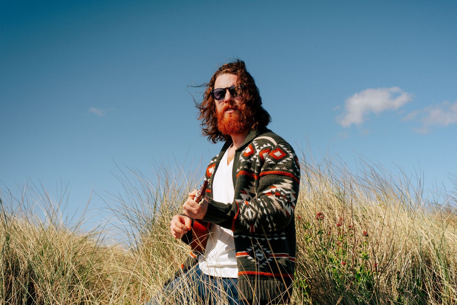 Someone wearing sunglasses and a patterned sweater stands in tall grass against a blue sky background. PHOTOGRAPHER Dublin
