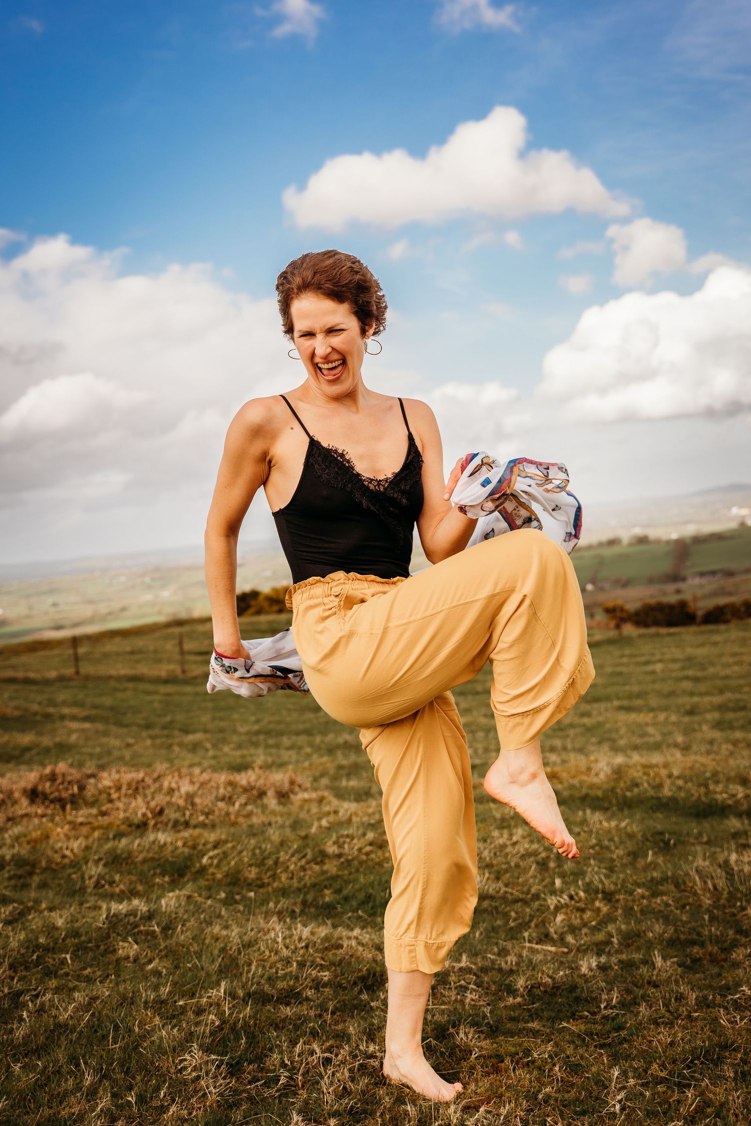 Person in a black tank top and yellow pants dances joyfully in a grassy field under blue sky with white clouds. Portrait PHOTOGRAPHER Dublin