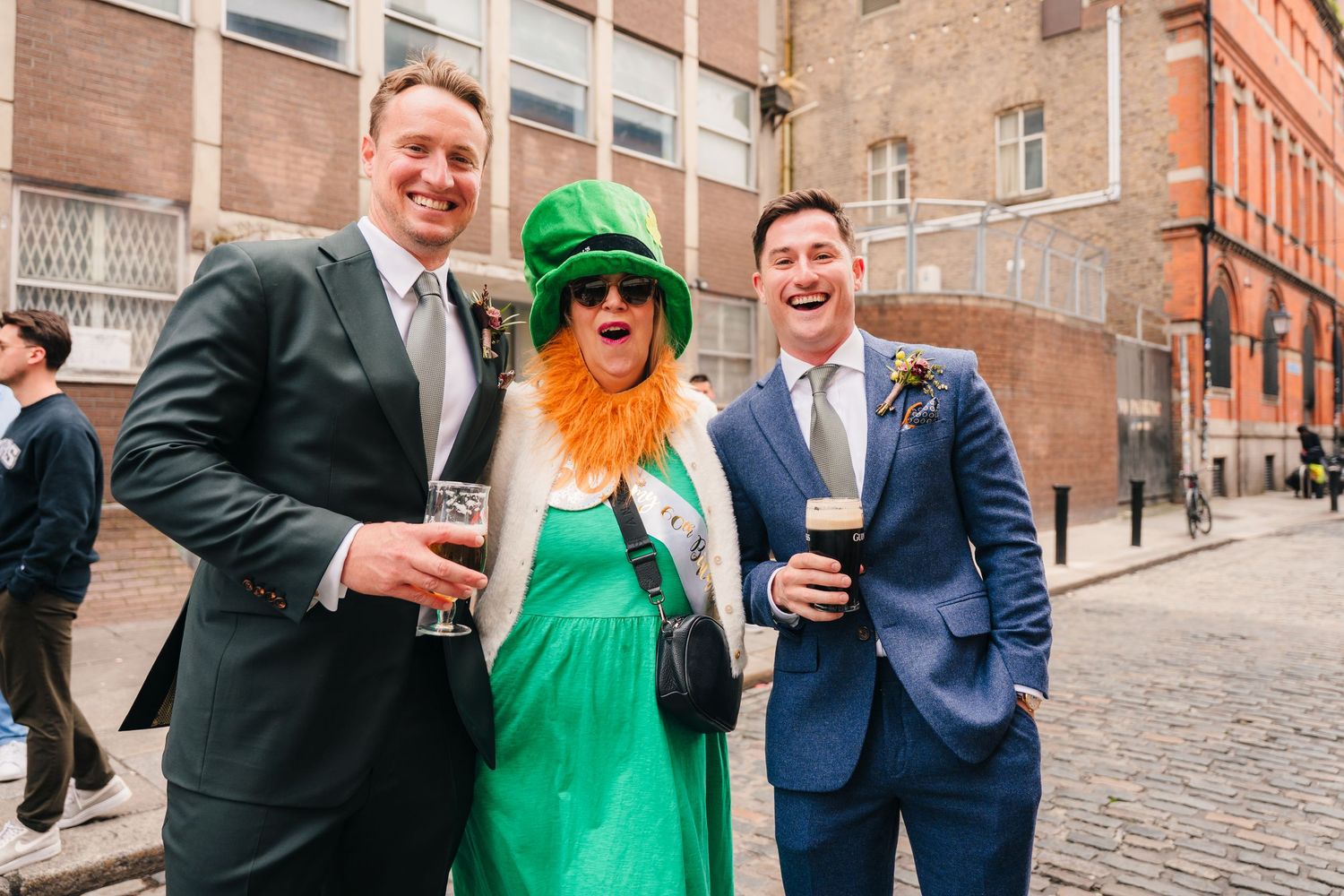Three happy wedding guests dressed in formal attire and St. Patrick's Day accessories pose on a cobblestone street. Same sex wedding PHOTOGR