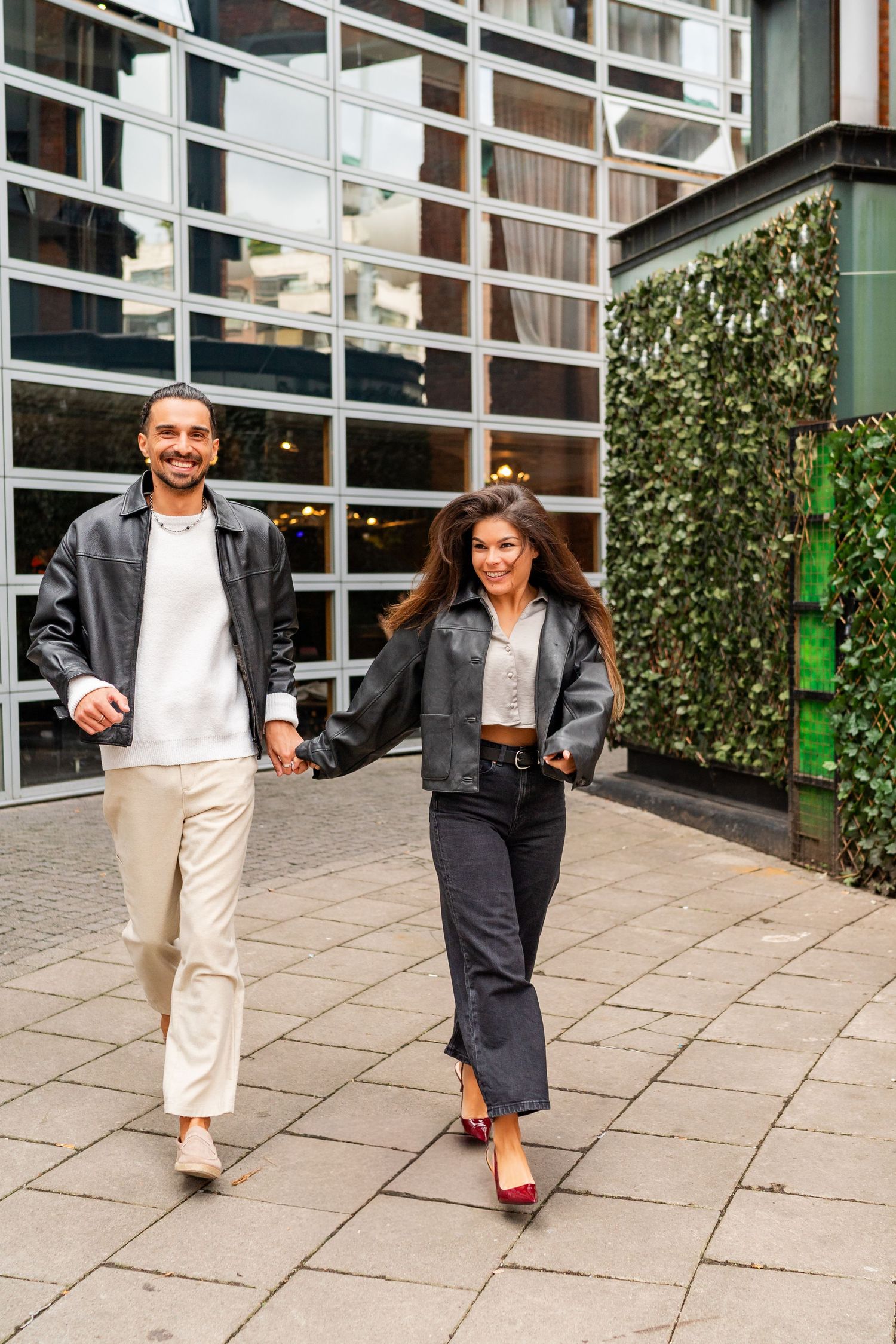 Couple walking and holding hands on a paved courtyard outside a modern glass building in matching casual outfits. Couple's Photoshoot PHOTOG