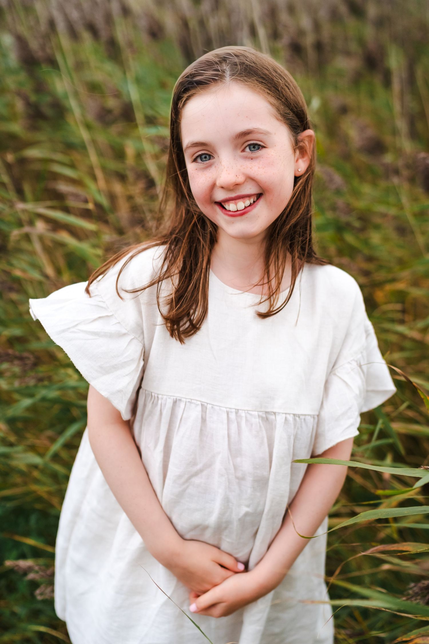 Young girl in a white dress smiling in front of tall grass.  Family photographer Dublin Ireland