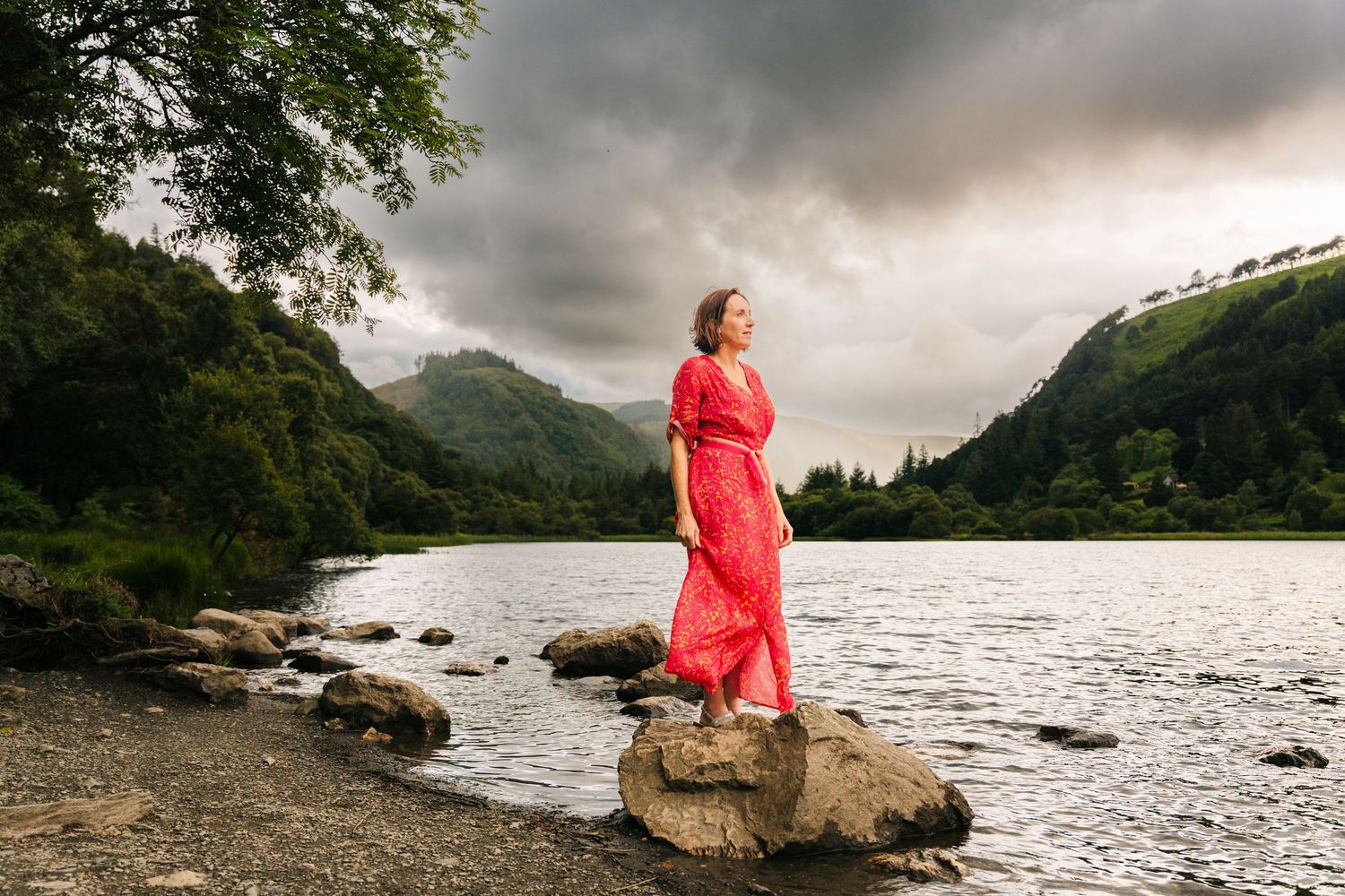 A figure in a red dress poses on rocks near a dramatic mountain lake under moody storm clouds. PHOTOGRAPHER Dublin ireland
