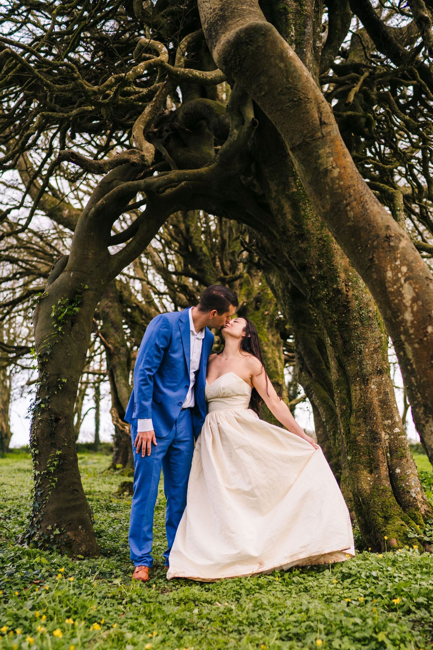 A couple in formal attire poses romantically under gnarled tree branches in a lush green setting. PHOTOGRAPHER Dublin ireland