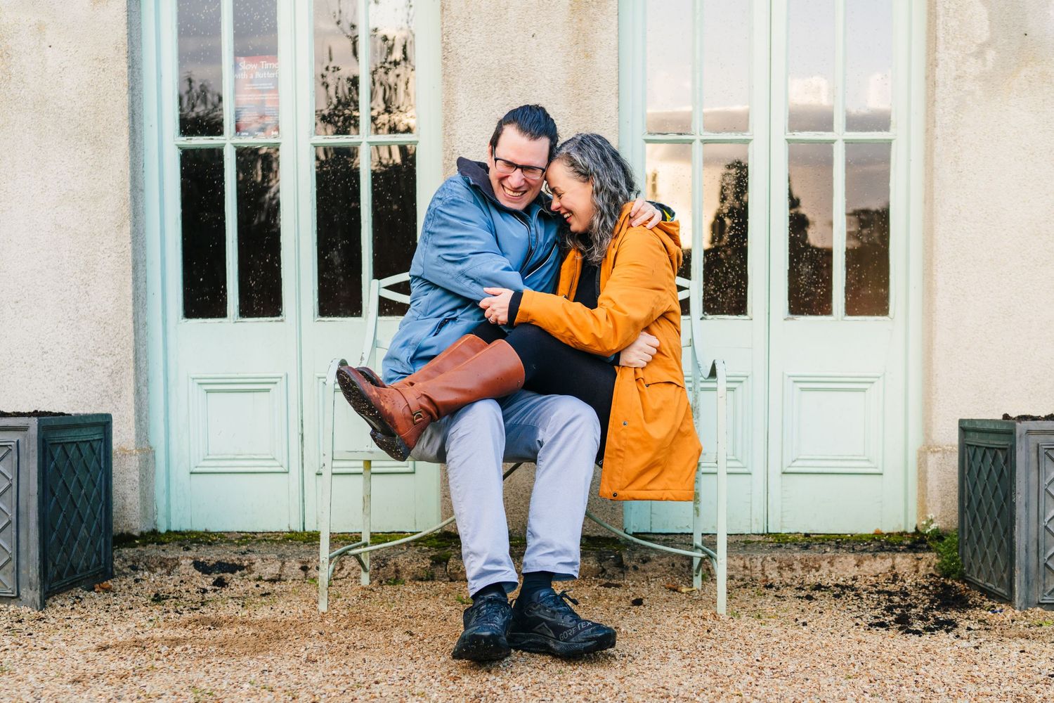 A couple sitting close together on a bench in front of mint green doors, wearing bright yellow and blue winter coats. PHOTOGRAPHER Dublin ir