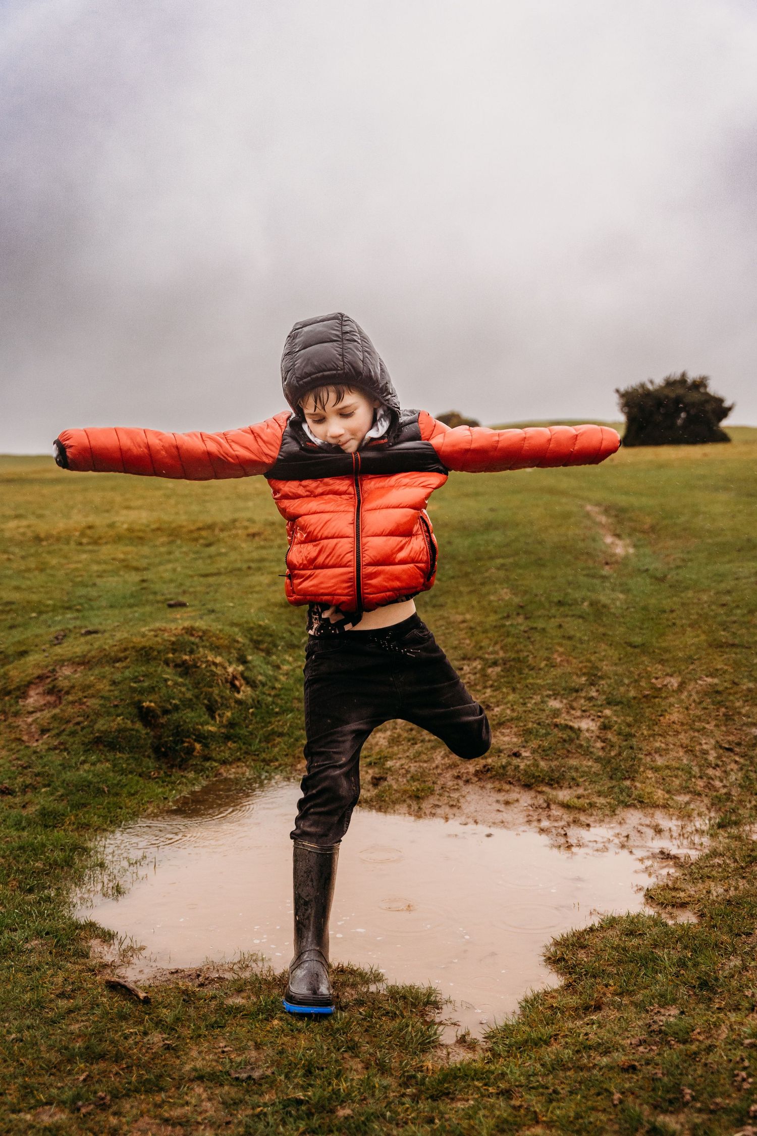 Small child wearing an orange puffy coat and winter boots plays in a puddle while spreading their arms on a cloudy day. family PHOTOGRAPHER