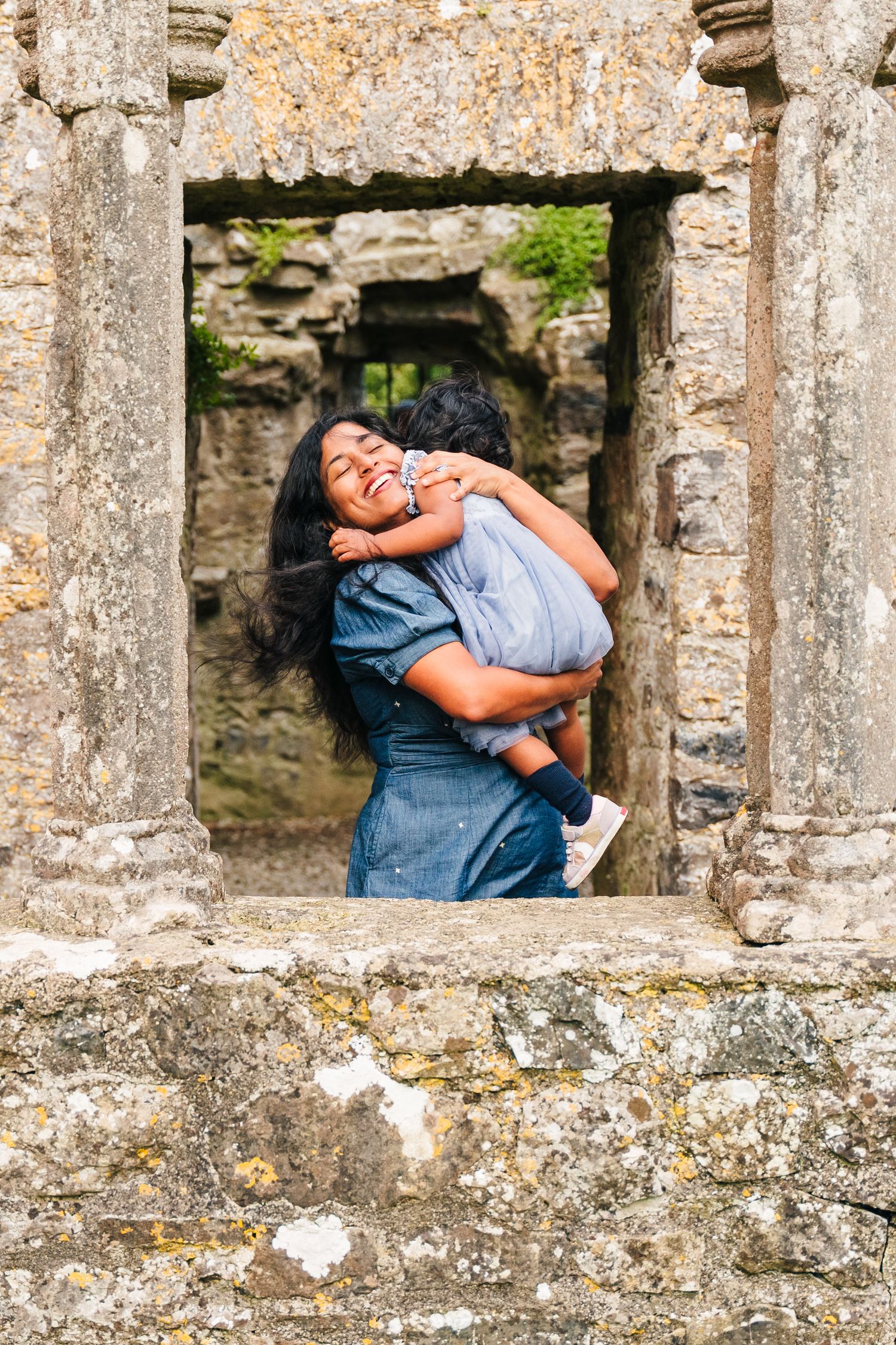 A tender moment captured through a stone window frame at an ancient ruins location, featuring denim and casual attire. family PHOTOGRAPHER D