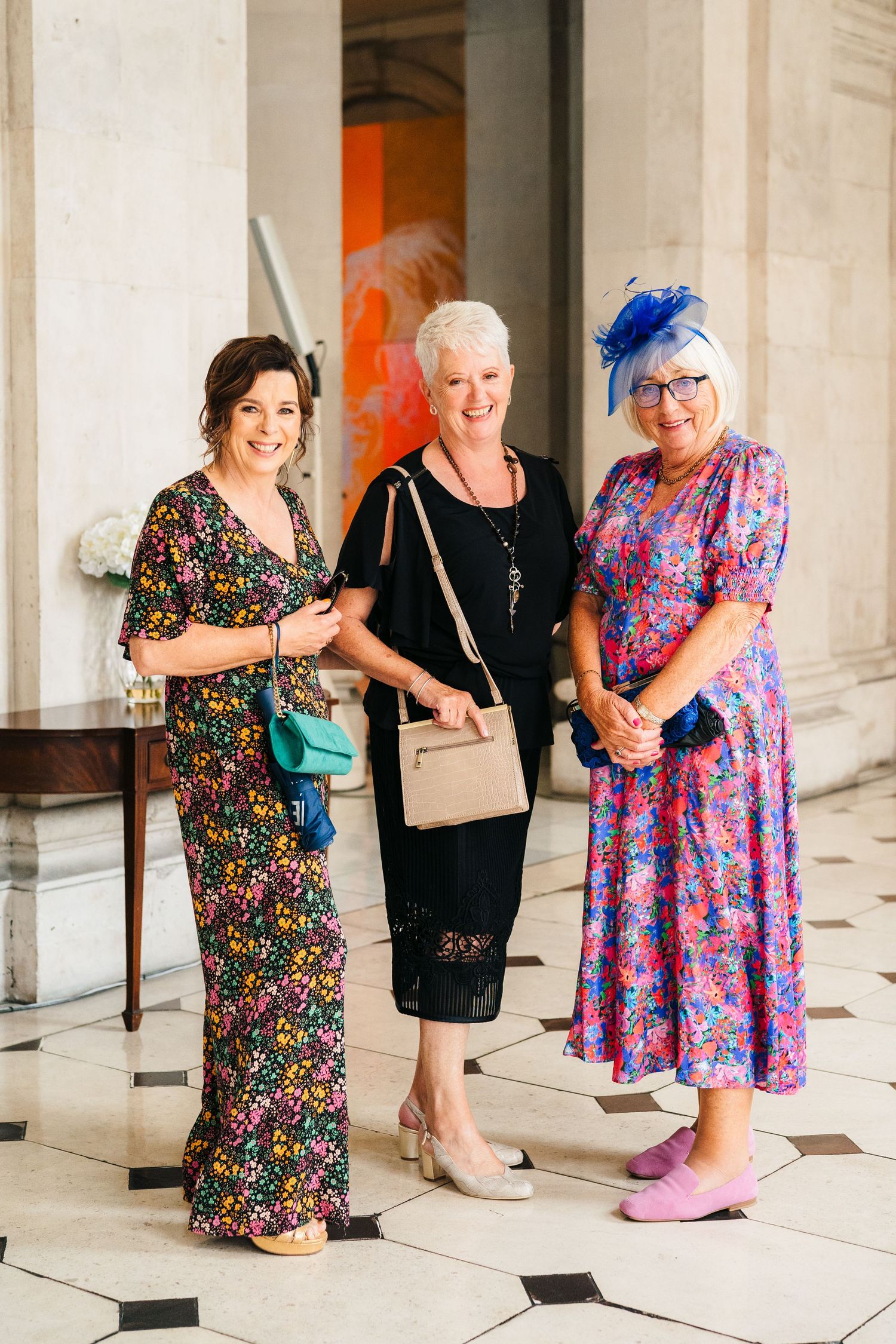Three wedding guests in colorful floral dresses and elegant hats pose together in an ornate hallway.  city hall dublin wedding photos