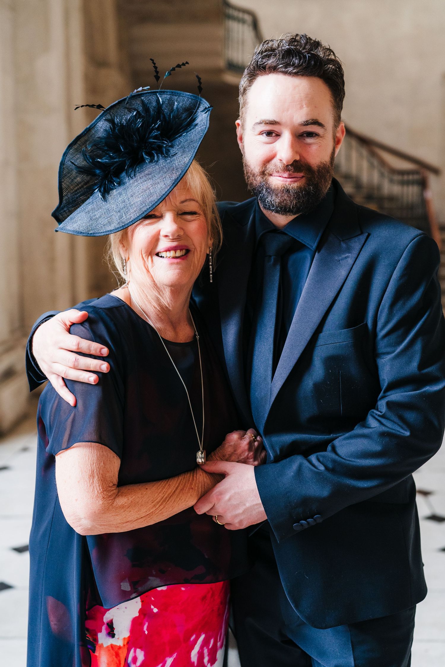 Formal event attendees wearing navy suit and black dress with striking blue fascinator pose in elegant venue.  city hall dublin wedding phot