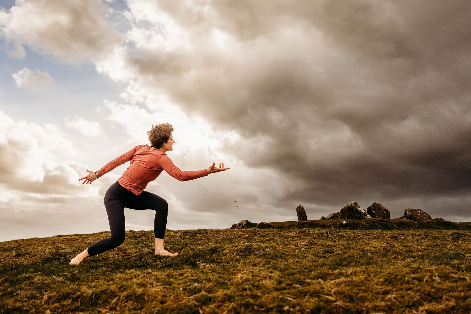 A person in an orange top strikes a dynamic pose while standing on a grassy hill under moody clouds. PHOTOGRAPHER Dublin ireland