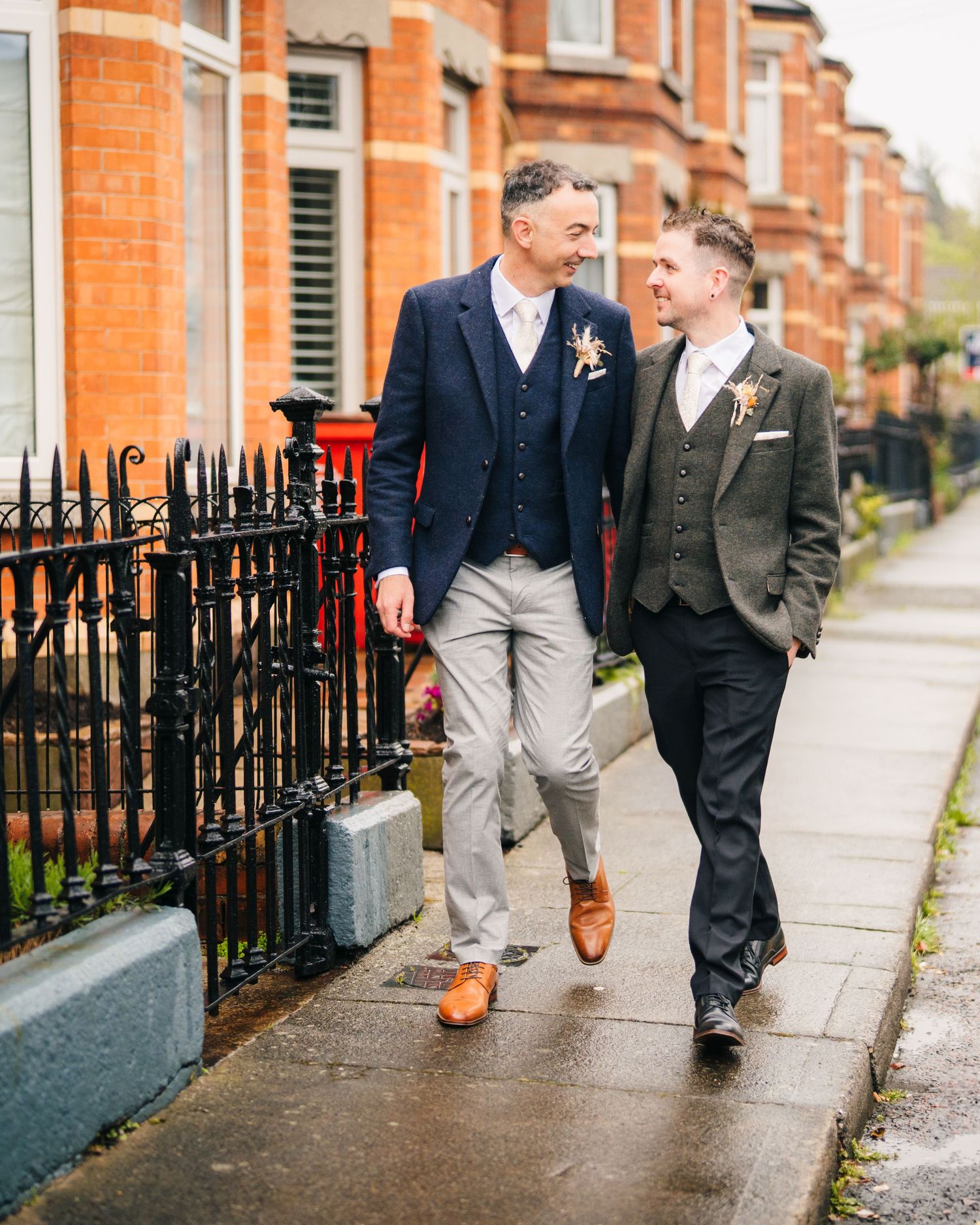 Two wedding guests in stylish suits walking along a brick-lined street with iron fencing in London. Marco Pierre Dublin Wedding Photographer