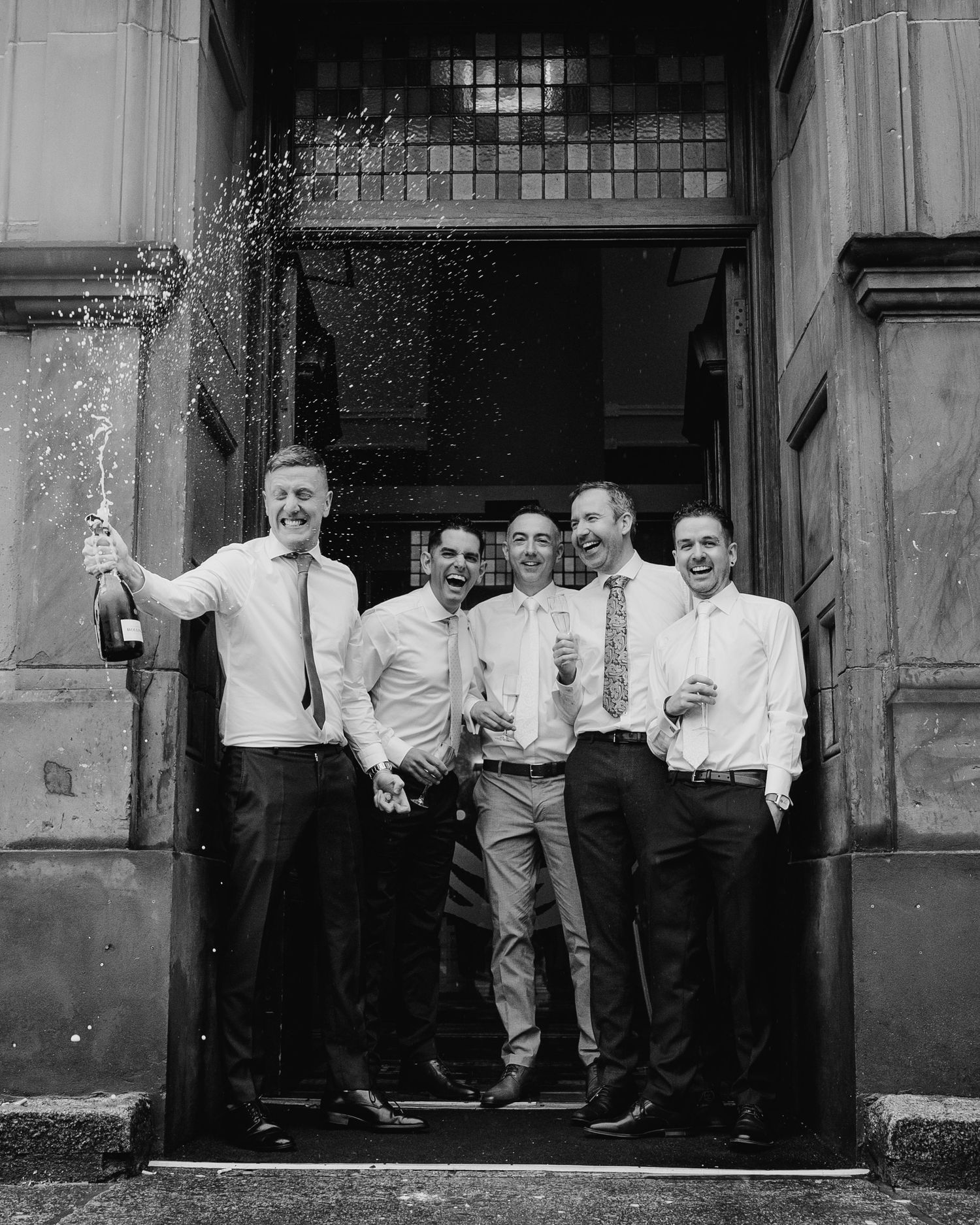 Black and white sequence showing groomsmen spraying champagne outside historic building entrance. Marco Pierre Dublin Wedding Photographer
