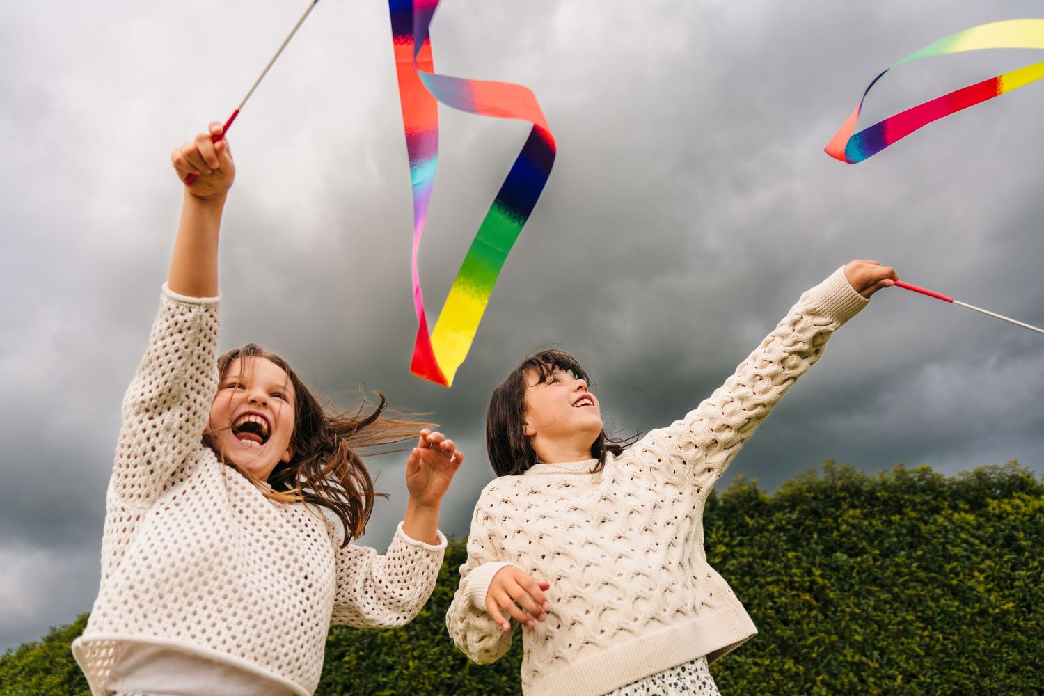 Two people in cream sweaters laugh and wave colorful ribbons against a dark cloudy sky with green hedges behind them. family PHOTOGRAPHER Du