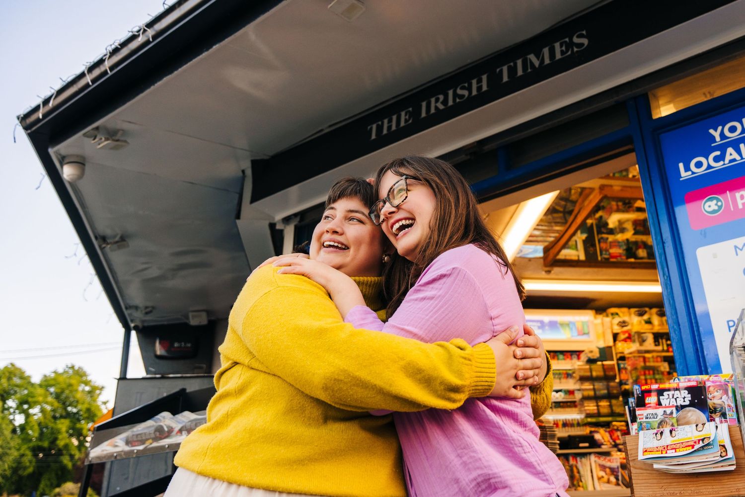 Two friends laughing and embracing while standing at an outdoor retail storefront. family PHOTOGRAPHER Dublin
