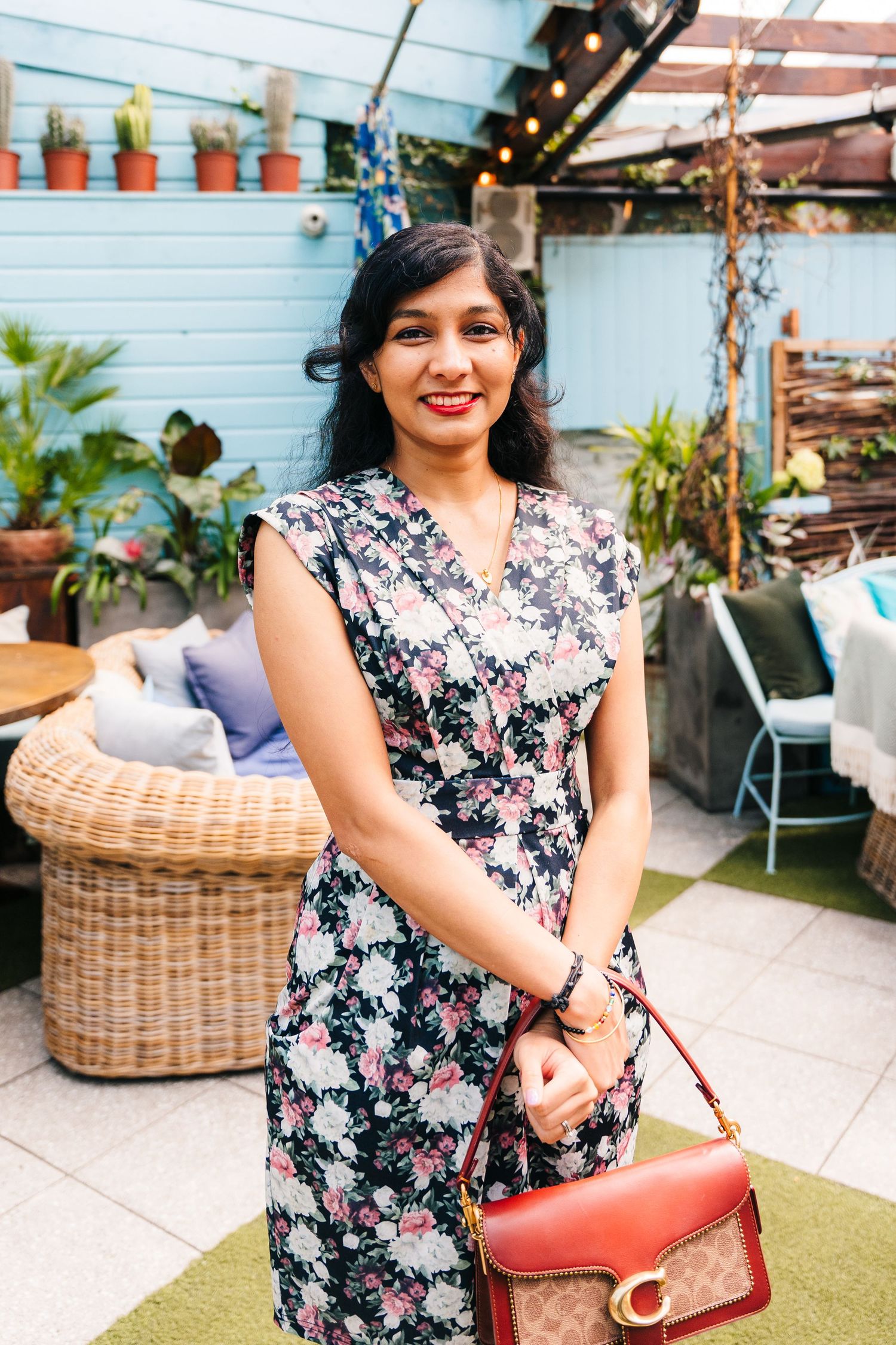 A woman in a floral print dress poses with a red handbag on an outdoor patio with wicker furniture and plants. House Dublin Wedding Photogra