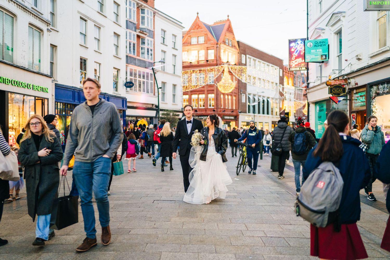 Bride and groom exploring busy city sidewalks among pedestrians and storefronts. Wedding PHOTOGRAPHER Dublin